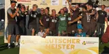 Hinter einem, von zwei Kindern gehaltenen Schild mit der Aufschrift "Deutscher Meister" steht das FC St. Pauli-Team in braunen Trikots und jubelt in die Kamera. Hippo hält die Meisterschale