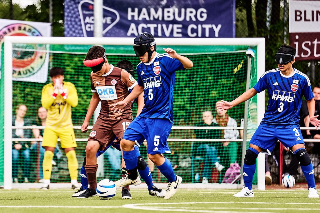 Mo Safari im Zweikampf um den Ball mit einem japanischen Nationalspieler. Im Hintergrund der japanische Torhüter und Banner am Zaun von Honey Badgers, Active City und FCSP Blindenfussball 